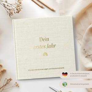 Op de afbeelding: Een wit boek met een linnen omslag, met gouden letters die "Dein erstes Jahr" (Je eerste jaar) en een gouden regenboog symbool lezen. Het boek heeft de tekst "Wundervolle Erinnerungen und Gl&uuml;cksmomente" (Prachtige herinneringen en gelukkige momenten) onderaan gedrukt. Het boek heeft ook een kleine Duitse vlag en een globe symbool met de tekst "In Deutschland designed, produziert & verpackt" (Ontworpen, geproduceerd en verpakt in Duitsland) en "Klimaneutral hergestellt" (Klima neutraal geproduceerd).