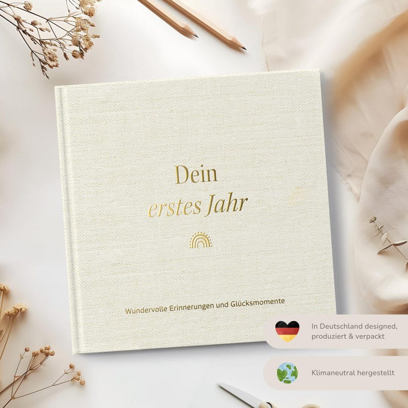 Op de afbeelding: Een wit boek met een linnen omslag, met gouden letters die "Dein erstes Jahr" (Je eerste jaar) en een gouden regenboog symbool lezen. Het boek heeft de tekst "Wundervolle Erinnerungen und Gl&uuml;cksmomente" (Prachtige herinneringen en gelukkige momenten) onderaan gedrukt. Het boek heeft ook een kleine Duitse vlag en een globe symbool met de tekst "In Deutschland designed, produziert & verpackt" (Ontworpen, geproduceerd en verpakt in Duitsland) en "Klimaneutral hergestellt" (Klima neutraal geproduceerd).