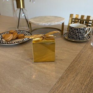 May include: A gold gift box with a tied ribbon sits on a table. A black and white patterned teacup and saucer are to the right. A plate of cookies and a marble cake stand are also on the table. A vase of white flowers is in the background.