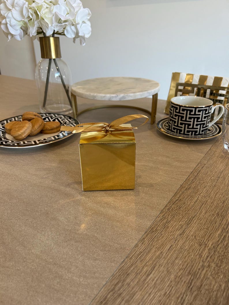 May include: A gold gift box with a tied ribbon sits on a table. A black and white patterned teacup and saucer are to the right. A plate of cookies and a marble cake stand are also on the table. A vase of white flowers is in the background.
