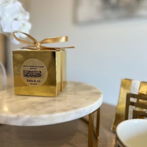 May include: A gold gift box with a gold ribbon sits on a white marble cake stand with gold legs. The box has a gold sticker with text in Arabic and English. A white floral arrangement is in the background.