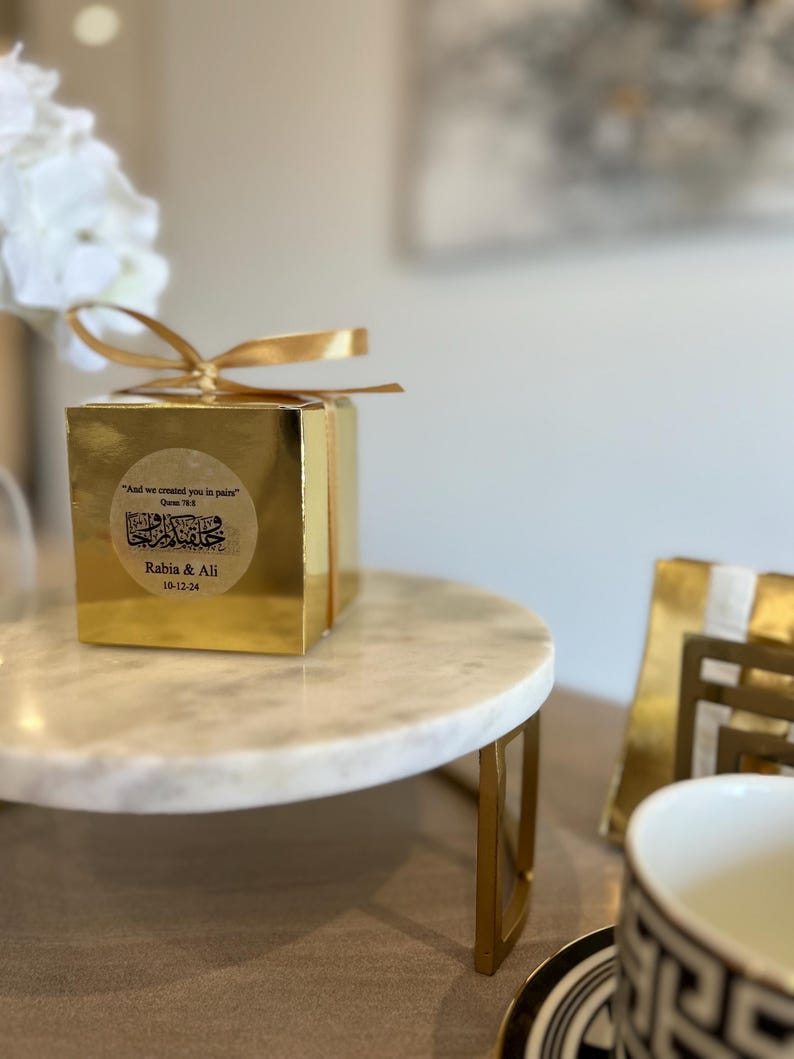 May include: A gold gift box with a gold ribbon sits on a white marble cake stand with gold legs. The box has a gold sticker with text in Arabic and English. A white floral arrangement is in the background.
