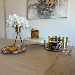 May include: A table setting featuring a clear glass vase with white flowers, a marble cake stand, a black and white patterned teacup and saucer, a clear glass, and a plate with cookies. A gold-framed abstract painting hangs in the background.