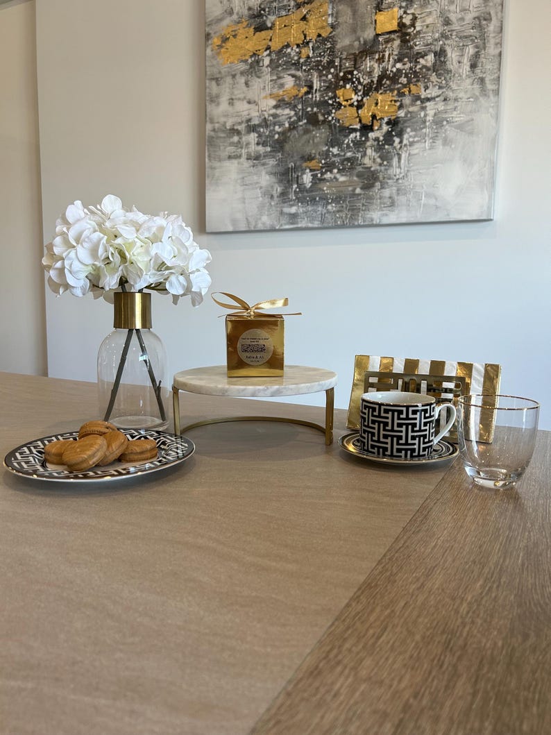 May include: A table setting featuring a clear glass vase with white flowers, a marble cake stand, a black and white patterned teacup and saucer, a clear glass, and a plate with cookies. A gold-framed abstract painting hangs in the background.