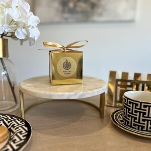 May include: A gold gift box with a gold ribbon sits on a marble cake stand with gold legs. A black and white patterned teacup and saucer are on the right. A vase with white flowers is on the left.