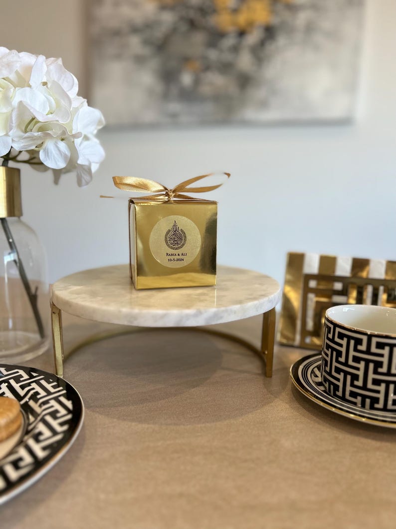 May include: A gold gift box with a gold ribbon sits on a marble cake stand with gold legs. A black and white patterned teacup and saucer are on the right. A vase with white flowers is on the left.