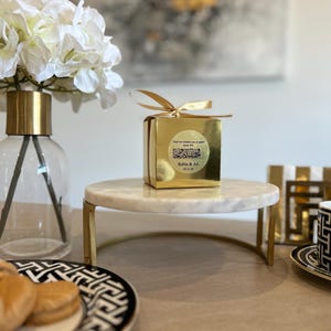 May include: A gold gift box with a gold ribbon sits on a white marble cake stand with gold legs. The box has text and a circular sticker. A vase of white flowers and a plate with pastries are also visible.
