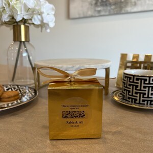 May include: A gold gift box tied with a gold ribbon, featuring a sticker with Arabic script and the text "Rabia & Ali 10-12-24". The box is on a table with a decorative plate, a marble stand, and a vase with white flowers.