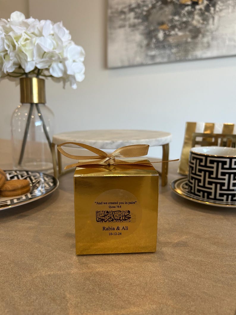 May include: A gold gift box tied with a gold ribbon, featuring a sticker with Arabic script and the text "Rabia & Ali 10-12-24". The box is on a table with a decorative plate, a marble stand, and a vase with white flowers.