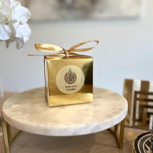 May include: A gold-colored gift box with a matching ribbon bow, sitting on a marble cake stand. The box has a circular sticker with Arabic text and the names "Rabia & Ali" and the date "10-5-2024".