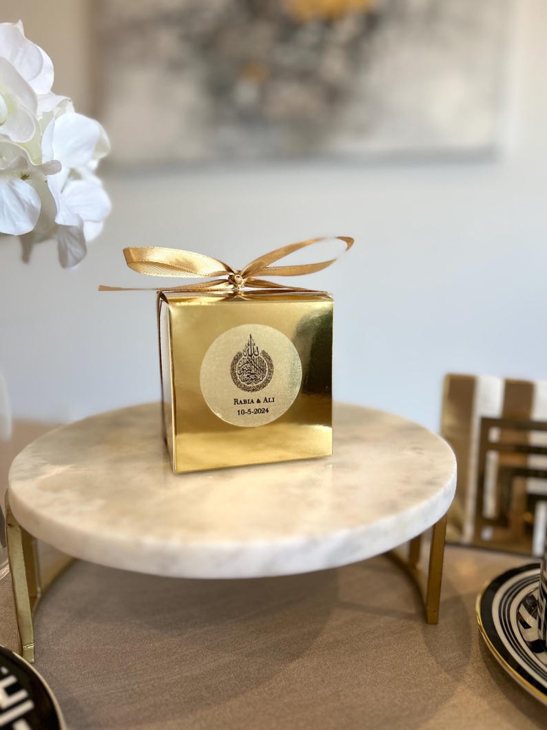 May include: A gold-colored gift box with a matching ribbon bow, sitting on a marble cake stand. The box has a circular sticker with Arabic text and the names "Rabia & Ali" and the date "10-5-2024".