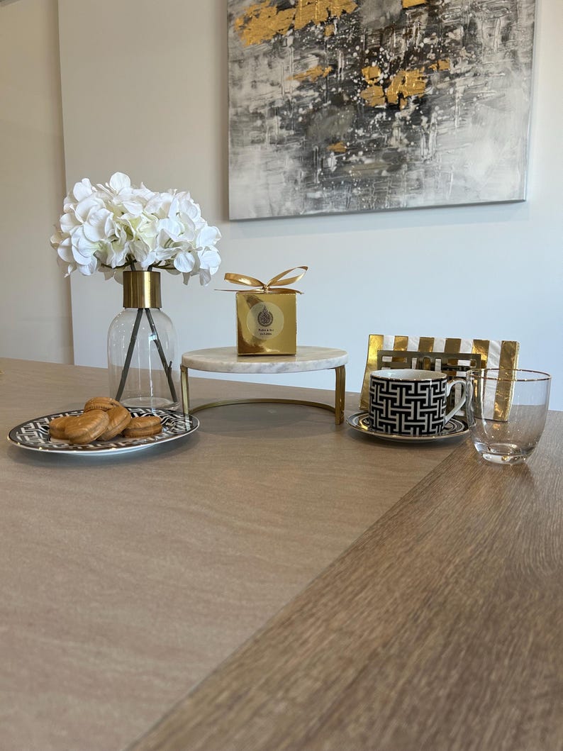 May include: A table setting featuring a clear glass vase with white flowers, a gold-accented box, a black and white patterned teacup, and a plate of cookies. A modern abstract painting hangs in the background.
