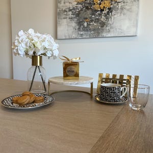 May include: A table setting featuring a clear glass vase with white flowers, a marble cake stand, a black and white patterned teacup and saucer, and a clear drinking glass. A plate of cookies and a gold-wrapped box are also present.