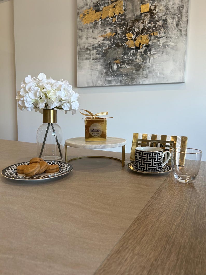 May include: A table setting featuring a clear glass vase with white flowers, a marble cake stand, a black and white patterned teacup and saucer, and a clear drinking glass. A plate of cookies and a gold-wrapped box are also present.