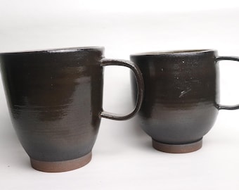 May include: Two dark brown ceramic mugs with rounded bottoms and handles. The mugs are made of a textured clay and have a glossy finish.