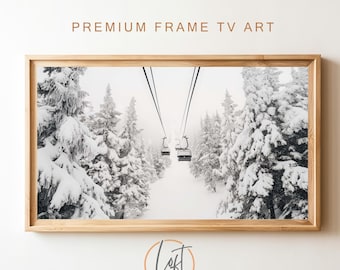 May include: A rectangular framed wall art piece featuring a black and white photograph of ski lift gondolas suspended over a snowy forest. The light-colored wooden frame surrounds the landscape image, which shows snow-covered pine trees.