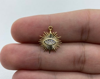 Puede incluir: Un dije dorado con un diseño de rayos de sol y un ojo en el centro. El ojo está detallado con pequeñas piedras claras y azules. El dije tiene un pequeño lazo en la parte superior para sujetarlo a la joyería.
