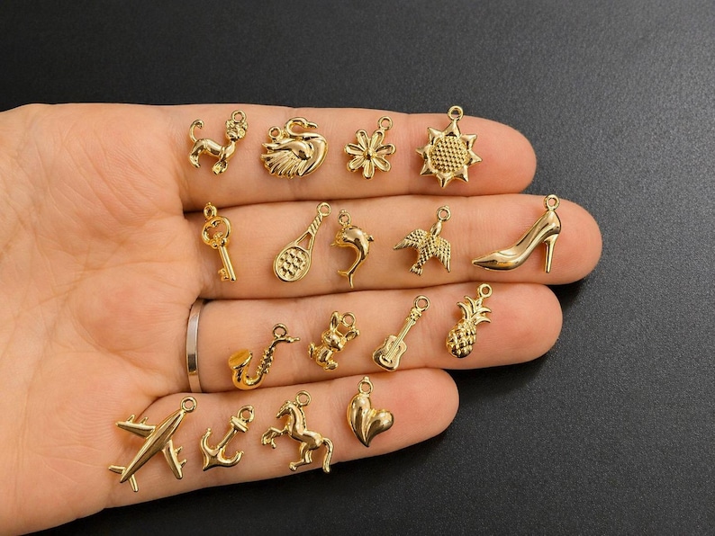 Puede incluir: Una colecci&oacute;n de peque&ntilde;os colgantes dorados de diversas formas: perro, cisne, llave, raqueta de tenis, delf&iacute;n, p&aacute;jaro, zapato de tac&oacute;n, saxof&oacute;n, guitarra, pi&ntilde;a, avi&oacute;n, ancla, caballo y coraz&oacute;n. Los colgantes se muestran sobre un fondo oscuro.