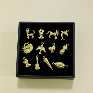 Puede incluir: Una colecci&oacute;n de dijes dorados en una caja negra. Los dijes incluyen varias formas como un le&oacute;n, un oso, un caballo, un delf&iacute;n, un cisne, un p&aacute;jaro, un coraz&oacute;n, una pi&ntilde;a, un oso de peluche, una guitarra, un cono y una concha. Los dijes son probablemente para hacer joyas.