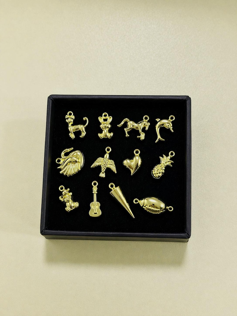 Puede incluir: Una colecci&oacute;n de dijes dorados en una caja negra. Los dijes incluyen varias formas como un le&oacute;n, un oso, un caballo, un delf&iacute;n, un cisne, un p&aacute;jaro, un coraz&oacute;n, una pi&ntilde;a, un oso de peluche, una guitarra, un cono y una concha. Los dijes son probablemente para hacer joyas.