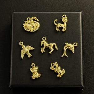 Puede incluir: Una colecci&oacute;n de dijes dorados con formas de animales, incluyendo un cisne, un gato, un p&aacute;jaro, un caballo, un delf&iacute;n, un oso y un conejo. Los dijes se muestran sobre una superficie negra, cada uno con un peque&ntilde;o lazo.
