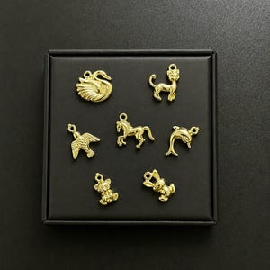 Puede incluir: Colecci&oacute;n de dijes dorados con formas de animales, incluyendo un cisne, un gato, un p&aacute;jaro, un caballo, un delf&iacute;n, un oso de peluche y un conejo. Los dijes se muestran sobre una superficie negra, cada uno con un lazo.