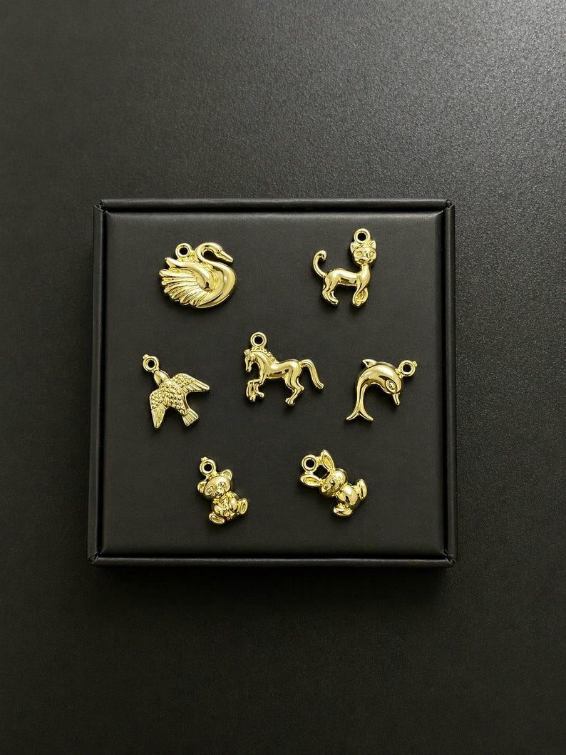 Puede incluir: Colecci&oacute;n de dijes dorados con formas de animales, incluyendo un cisne, un gato, un p&aacute;jaro, un caballo, un delf&iacute;n, un oso de peluche y un conejo. Los dijes se muestran sobre una superficie negra, cada uno con un lazo.