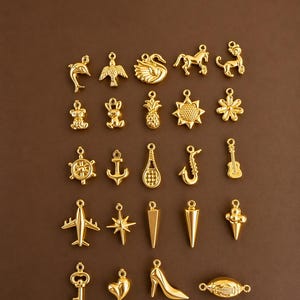 Puede incluir: Una colecci&oacute;n de colgantes dorados de varias formas, incluyendo un delf&iacute;n, un p&aacute;jaro, un cisne, un caballo, un mono, un oso de peluche, una pi&ntilde;a, una flor, un tim&oacute;n, un ancla, una raqueta de tenis, un saxof&oacute;n, una guitarra, un avi&oacute;n, una estrella, etc.