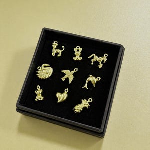 Puede incluir: Una colecci&oacute;n de dijes dorados en una caja negra. Los dijes incluyen un perro, un oso, un caballo, un cisne, un p&aacute;jaro, un delf&iacute;n, un coraz&oacute;n y una pi&ntilde;a. Los dijes son peque&ntilde;os y detallados, con un lazo en la parte superior para sujetarlos.