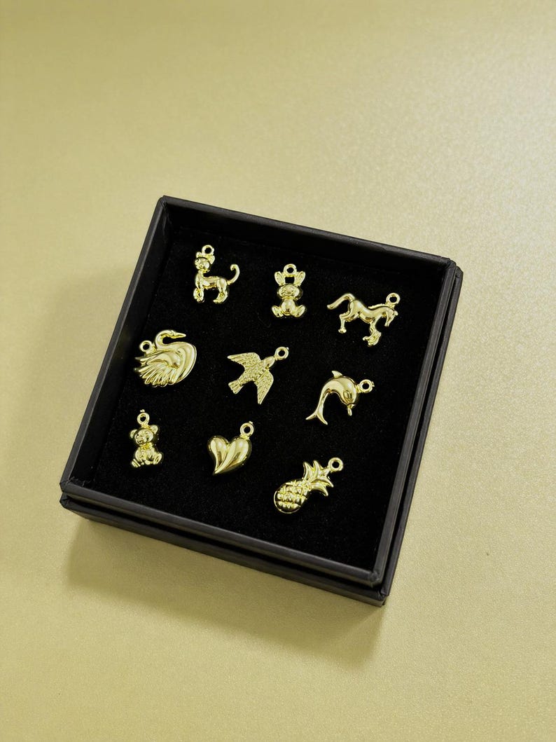 Puede incluir: Una colecci&oacute;n de dijes dorados en una caja negra. Los dijes incluyen un perro, un oso, un caballo, un cisne, un p&aacute;jaro, un delf&iacute;n, un coraz&oacute;n y una pi&ntilde;a. Los dijes son peque&ntilde;os y detallados, con un lazo en la parte superior para sujetarlos.
