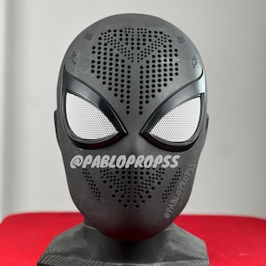 May include: Black Spider-Man mask with a black and white spider design and a black base. The mask has a textured surface with small holes throughout. The mask is on a black stand. The text "@PABLOPROPSSS" is visible on the mask.