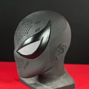 May include: A black Spider-Man mask with a textured surface and a sleek, angular design. The mask features white mesh eye coverings and a distinctive, pointed eye shape. The mask is displayed on a gray stand against a red background.