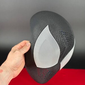 May include: A black mask with a textured surface featuring large, white, mesh-covered eye openings. The mask has a curved, contoured shape with a pattern of small, circular perforations across the forehead and cheek areas, providing a detailed look.