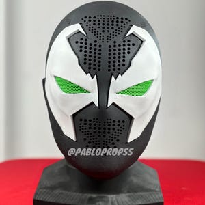 Puede incluir: Una máscara impresa en 3D en blanco y negro con ojos verdes. La máscara está en un soporte negro. El texto "@PABLOPROPSSS" está impreso en la máscara.