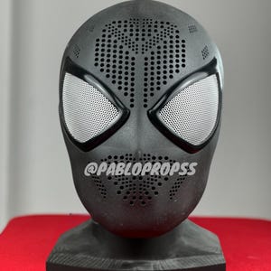 Op de afbeelding: Een zwart Spider-Man-masker met witte mesh oogbedekkingen en een geperforeerd ontwerp op het voorhoofd en de mond. Het masker wordt getoond op een zwarte standaard tegen een rode achtergrond. De tekst "@PABLOPROPS" is zichtbaar.
