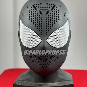 May include: Black Spider-Man mask with a white spider logo and white mesh eyes. The mask is on a black stand and has a textured surface. The text "@PABLOPROPSS" is printed on the mask.