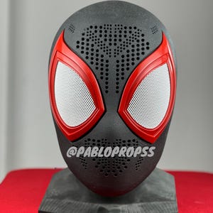 May include: A black and red Spider-Man mask with a white mesh eye piece. The mask has a textured surface with small holes. The mask is on a black stand.