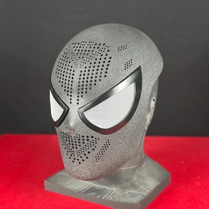 Op de afbeelding: Een getextureerd, donkergrijs Spider-Man-masker met zwarte accenten en witte mesh oogbedekkingen. Het masker heeft talrijke kleine perforaties en wordt gepresenteerd op een grijze standaard, tegen een rode achtergrond.