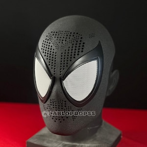 Op de afbeelding: Een zwart Spider-Man-masker met witte mesh oogbedekkingen en een geperforeerd ontwerp op het voorhoofd. Het masker wordt weergegeven op een grijze standaard tegen een rode en zwarte achtergrond. De tekst "@PABLOPROPS" is zichtbaar op het masker.