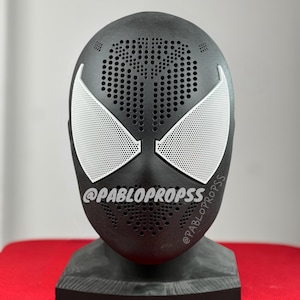 May include: A black Spider-Man mask with white eye lenses and a perforated design. The mask is displayed on a black stand. The text "@PABLOPROPS" is printed on the mask.