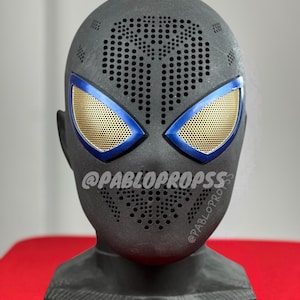 Op de afbeelding: Een donkergrijs masker met blauwe en gouden accenten, met een geperforeerd patroon over het voorhoofd en de mond. De oogopeningen zijn van goudkleurig gaas, omlijnd met blauw. De tekst "@PABLOPROPS" is zichtbaar op het masker.