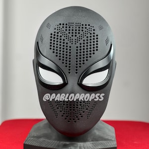 May include: A black superhero mask with white mesh eye coverings and a perforated design. The mask has a textured surface and is mounted on a gray base. The text "@PABLOPROPS" is printed on the mask.