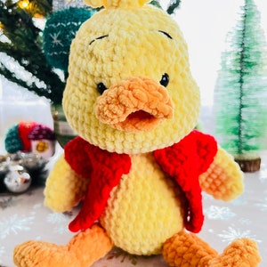May include: A crocheted yellow duck wearing a red vest. The duck has orange feet and a yellow beak. The duck is sitting on a white surface with a snowflake pattern.