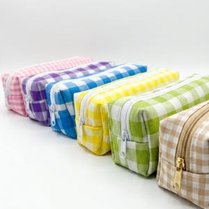Op de afbeelding: Een rij van zes rechthoekige etuis met rits in een vichy-ruitpatroon. De kleuren zijn roze, paars, blauw, geel, groen en beige. Elk etui heeft een witte rits, behalve het beige etui dat is voorzien van een goudkleurige metalen ritssluiting.