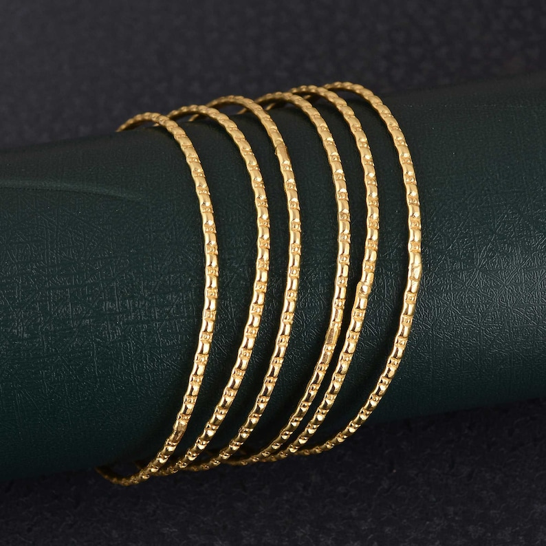 May include: A set of six gold bangles with a textured, linear design. The bangles are arranged in a curved line, showcasing their intricate pattern and warm, golden hue. The jewelry is displayed against a dark green background.