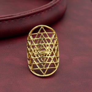 May include: A gold-colored ring with an intricate geometric design. The ring features a complex pattern of intersecting lines and shapes, creating a lattice-like appearance. The ring is set against a burgundy background.