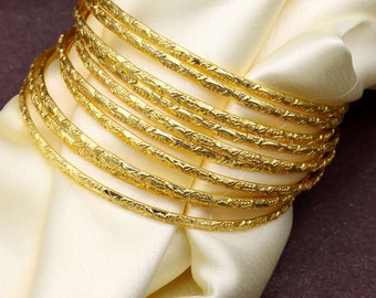 May include: A set of ten gold-toned bangle bracelets with an intricate floral design. The bracelets are stacked on top of each other and are lying on a white fabric surface.