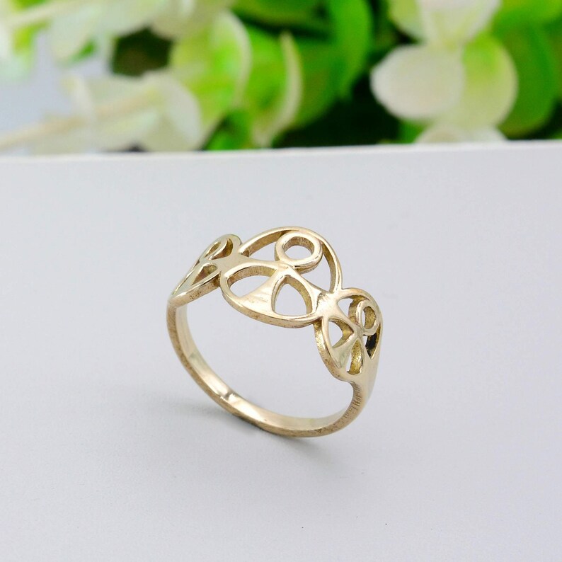 May include: A gold-colored ring with an openwork design. The ring features a series of interconnected, abstract shapes. The band is smooth and simple, while the top section showcases the decorative pattern. The ring is set against a soft, neutral background.