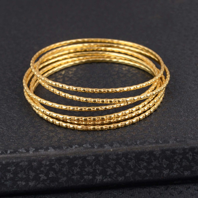 May include: A stack of six gold-colored bangles. Each bangle has a textured surface with a repeating pattern of small rectangular shapes. The bangles are stacked on top of each other, creating a layered effect.