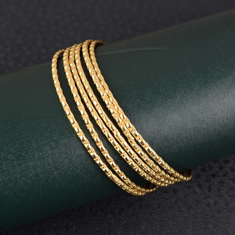 May include: A set of six gold bangles. Each bangle has a textured, repeating pattern. The bangles are stacked together, and are displayed on a dark green surface. The bangles are made of a shiny, gold material.